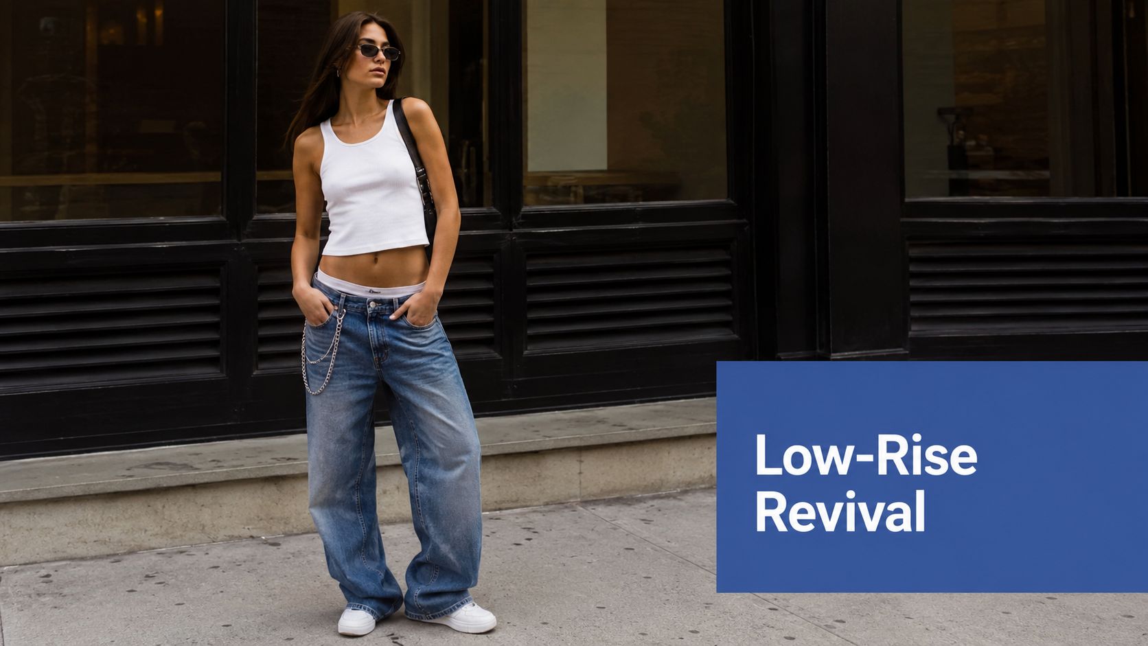 A young woman wearing a white tank top and baggy low-rise jeans standing on a city sidewalk.